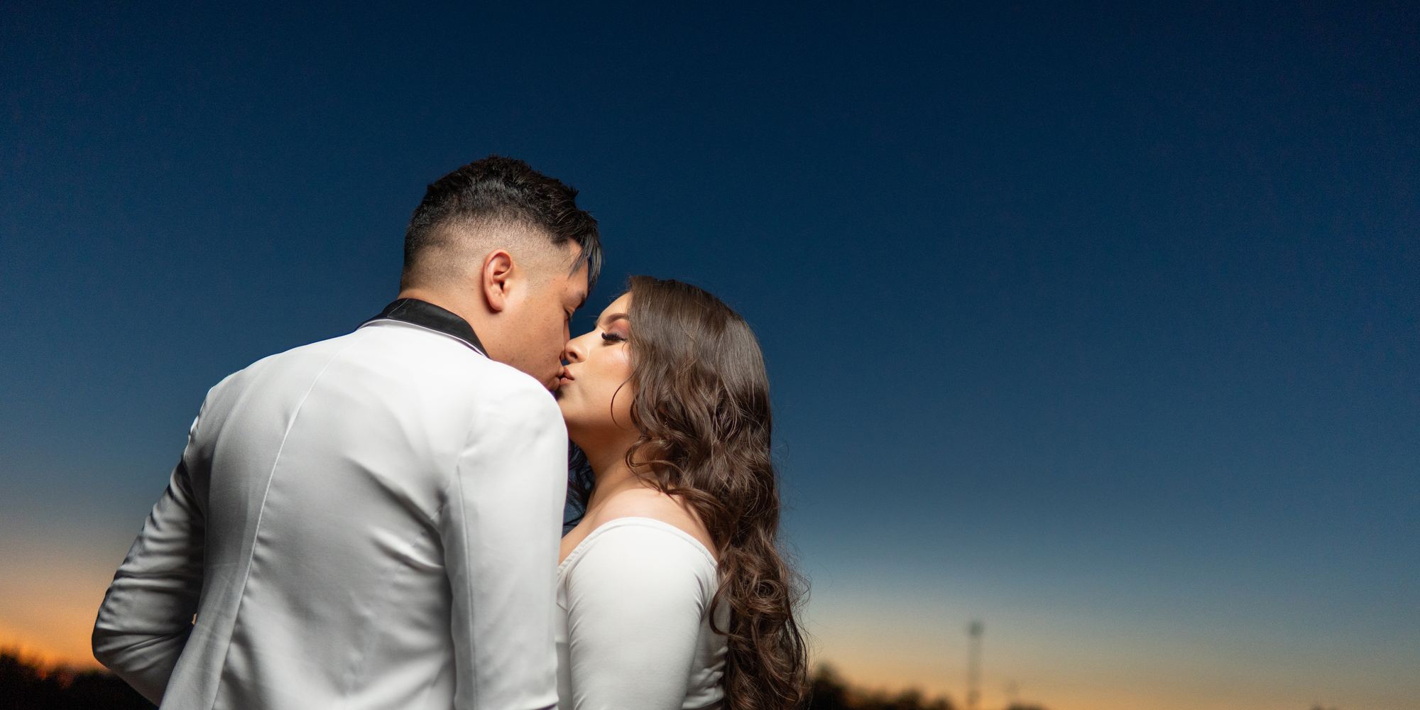 Novios besándose al atardecer bajo un cielo azul